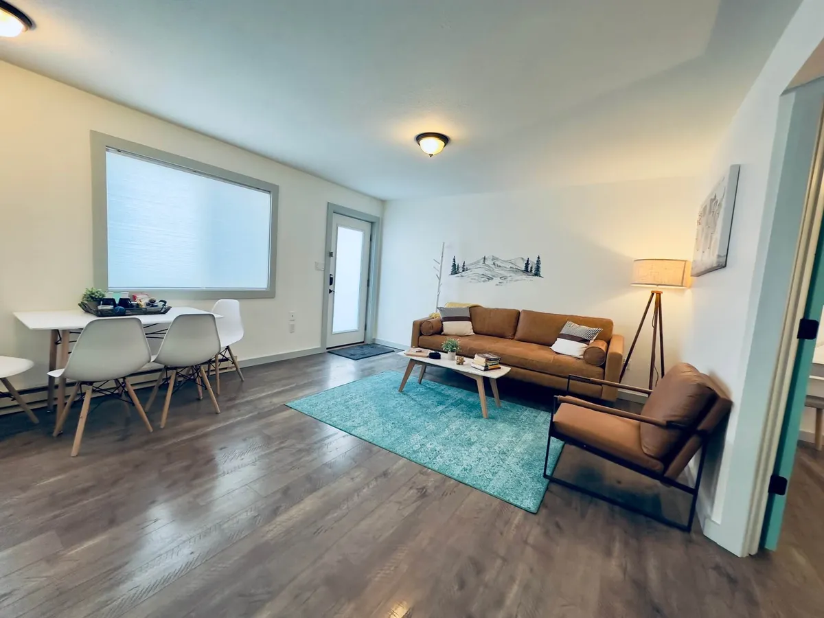 A welcoming living and dining area featuring a comfortable brown sofa, a dining table set for meals or games, a blue area rug adding a pop of color, warm lighting from a lamp, and tasteful wall art. The space feels open and inviting, ideal for gatherings and shared moments.