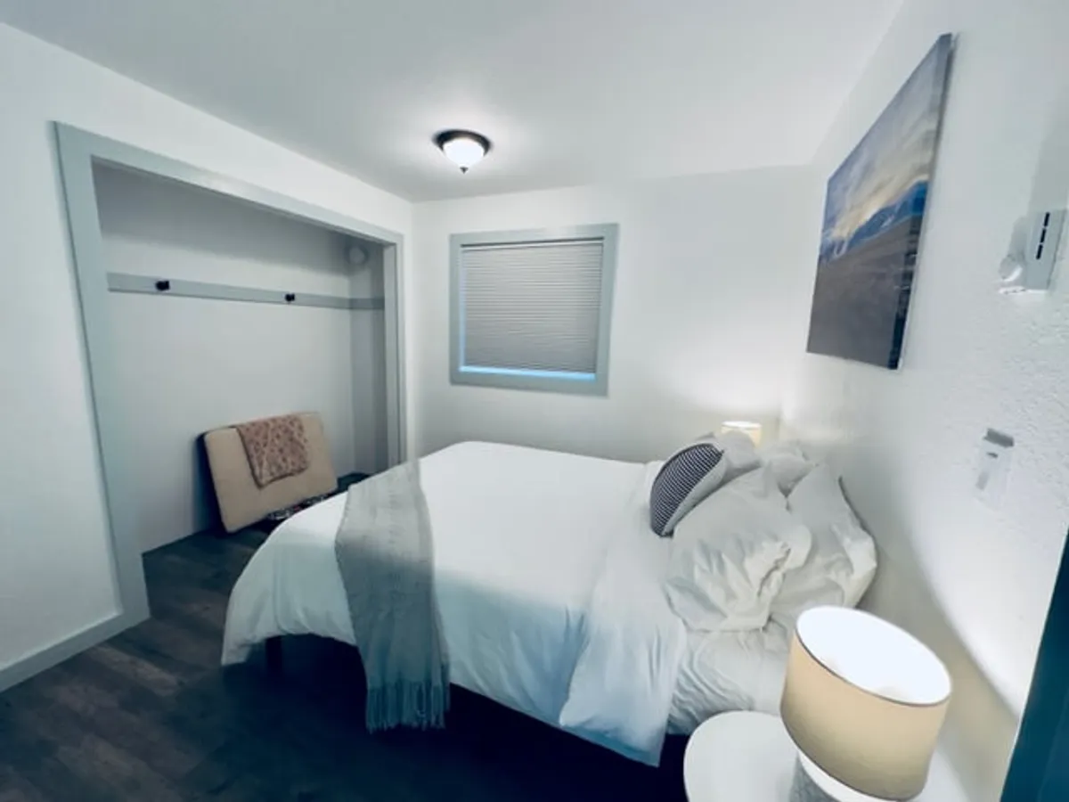 A cozy bedroom featuring a bed with crisp white linens, a decorative pillow, and a nightstand with a lamp. Artwork on the wall and a glimpse of the closet area create a lived-in, welcoming feel. The room is bathed in soft, natural light, inviting guests to relax and recharge.