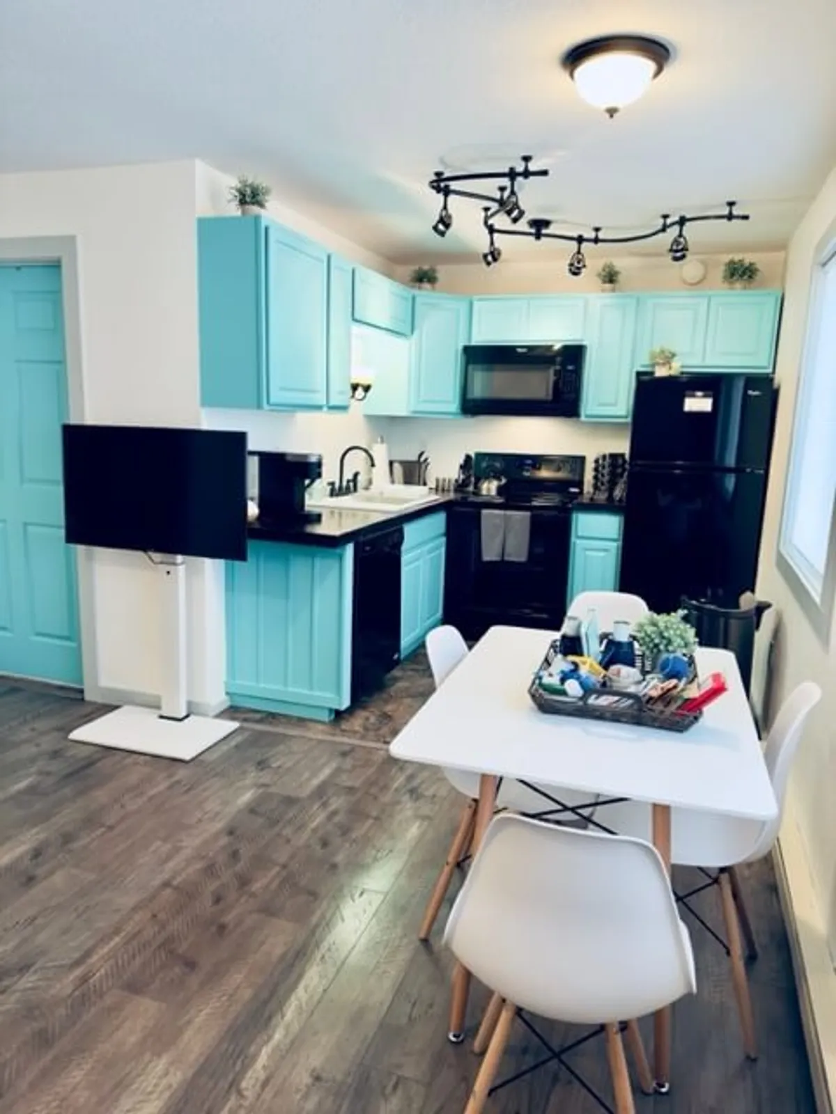 A bright, contemporary kitchen with standout blue cabinetry and a white dining table, all part of a spacious, open-plan layout. The scene is lively, showing guests gathered for cooking and socializing, with modern appliances visible, reinforcing the shared, communal cooking experience.