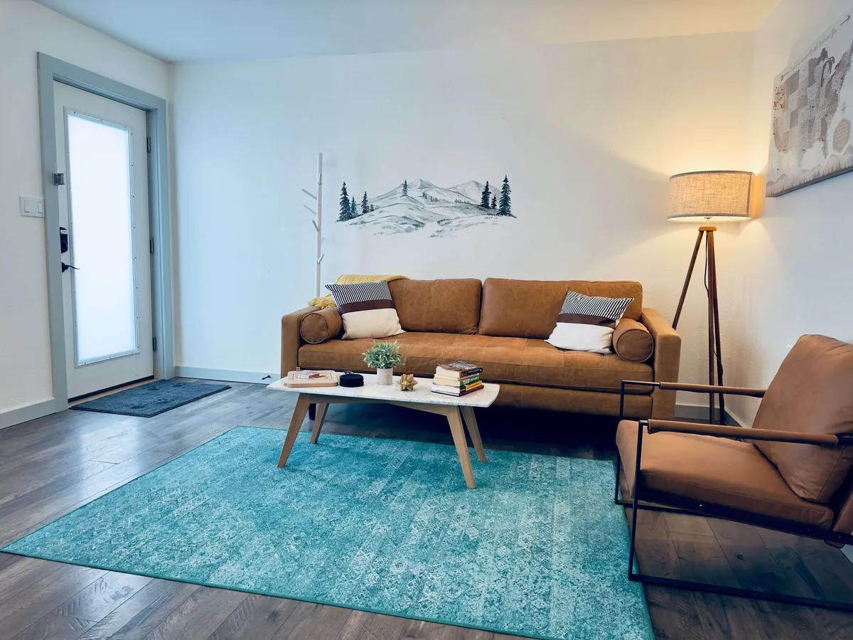 The image features a stylish living room interior with a cozy sofa, inviting coffee table, soft rug, warm floor lamp, and a striking mural of mountain scenery on the wall. The space is arranged for comfort, relaxation, and socializing, blending contemporary decor with natural inspiration.