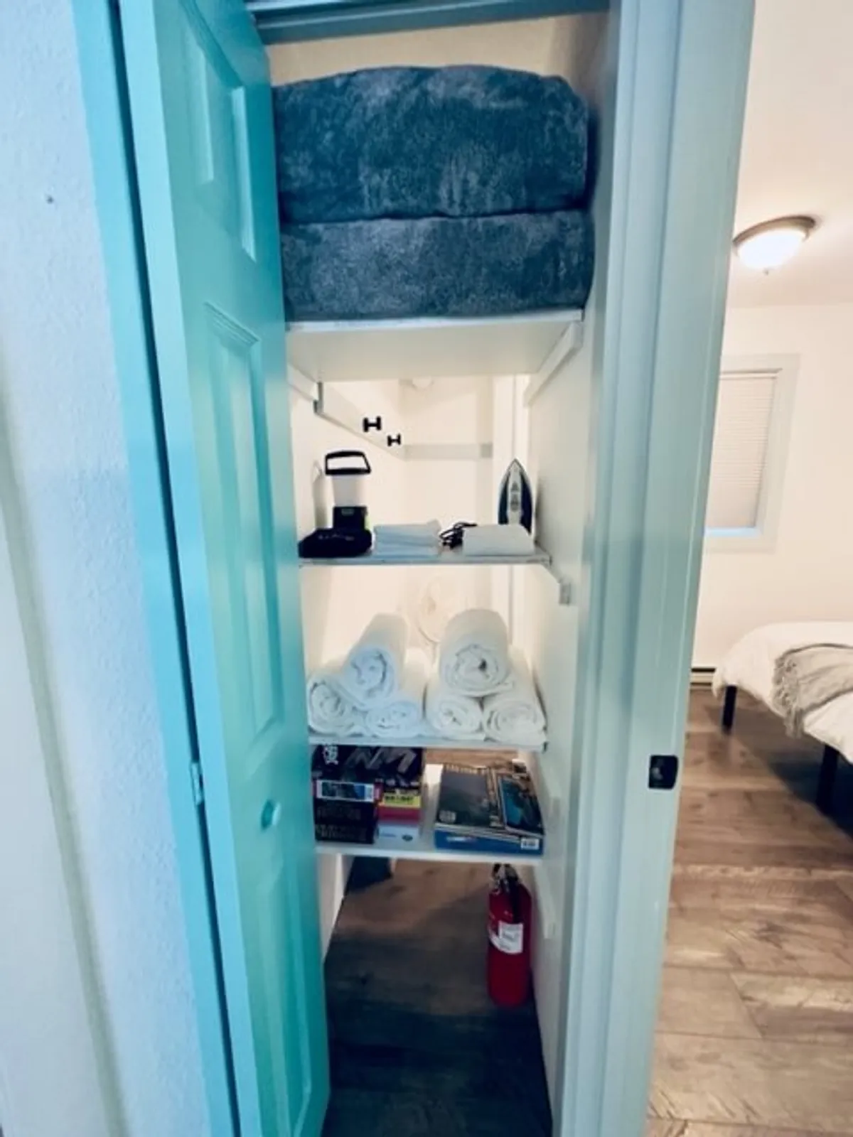 A functional nook with neatly stacked towels, shelves stocked with books and games, an iron and ironing board, and a fire extinguisher. The scene is practical yet inviting, emphasizing thoughtful touches that support a smooth and comfortable stay.